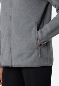 Persona che indossa una giacca in pile grigia con una tasca laterale con zip, mano nella tasca, abbinata a pantaloni neri.