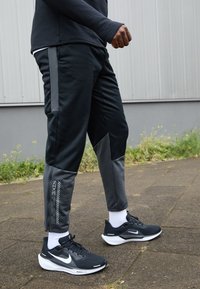 Osoba ubrana w czarne i szare spodnie sportowe oraz czarne buty do biegania Nike z białymi podeszwami, stojąca na utwardzonej powierzchni w pobliżu zieleni.