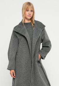 Grauer Wollmantel mit breiter Schulterklappe, langen Ärmeln und Seitentaschen. Strukturiertes Gewebe mit lockerem, oversize Schnitt.
