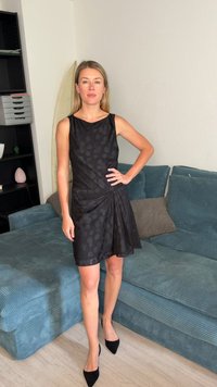 Femme en robe noire sans manches à motifs et talons noirs, debout, main sur la hanche dans le salon à côté d'un canapé bleu.