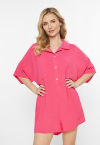 Rosa, kortärmad overall med krage, knapplist framtill, två fickor och en texturerad tygkvalitet. Avslappnad passform med sidoklipp för ökad rörelsefrihet.