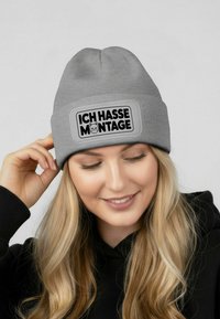 Gray ribbed beanie with a rectangular patch featuring the text "ICH HASSE MONTAGE" in bold black font and a cat graphic.