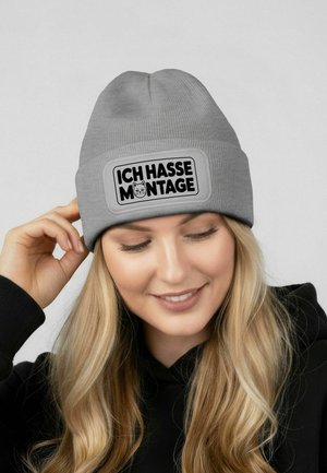 Gray ribbed beanie with a rectangular patch featuring the text "ICH HASSE MONTAGE" in bold black font and a cat graphic.
