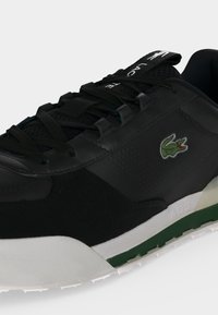 Μαύρο sneaker Lacoste με πράσινο λογότυπο κροκόδειλου, μαύρα κορδόνια και λευκή και πράσινη σόλα, ορατή από πλάγια γωνία.