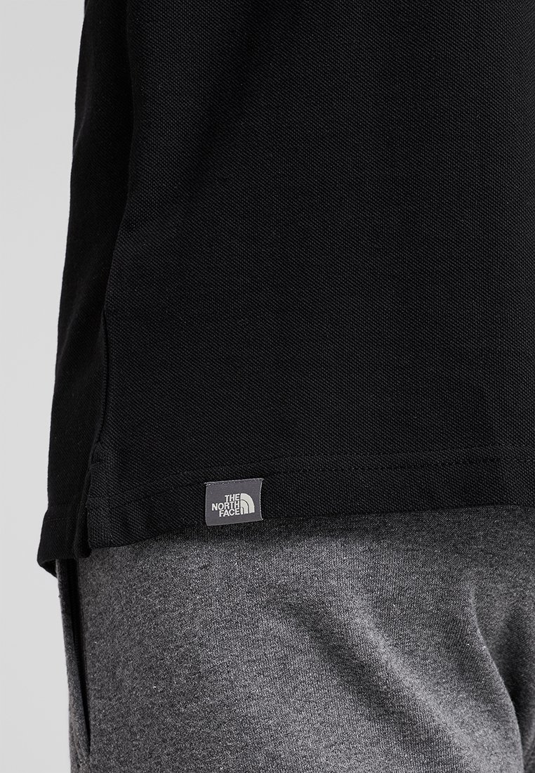 Černé tričko s krátkým rukávem z texturované tkaniny s viditelnou šedou logovou nášivkou "The North Face" na dolním lemu. Šedé kalhoty jsou částečně viditelné.