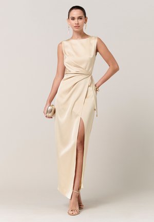 Mujer con un vestido de satén champán sin mangas y una abertura lateral, sosteniendo un bolso de mano a juego y luciendo pendientes y tacones dorados.