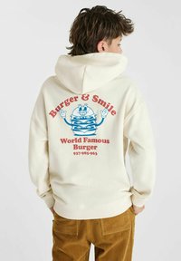 Creme-kleurige hoodie met op de rug een graphic van een cartoon hamburger en tekst in rood en blauw: "Burger & Smile, Wereldberoemde Hamburger."