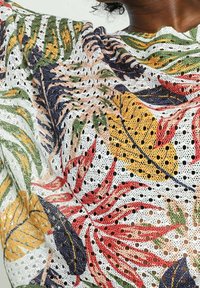 Blusa de encaje con un patrón de hojas multicolor sobre fondo blanco, con textura calada y mangas abullonadas.