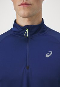Marineblauw sportieve quarter-zip top van een textuurstof, met een gele ritssluiting en een zilveren logo op de linkerkant van de borst.
