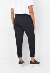 Person som bär svarta joggarbyxor med dragkedjor vid anklarna, vit t-shirt och svarta platåskor, stående mot en vit bakgrund.