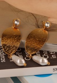 Gouden oorbellen met een geborsteld ovaal ontwerp, een heldere edelsteenaccent en een witte parel detail. Gevoeliger en visueel opvallend.