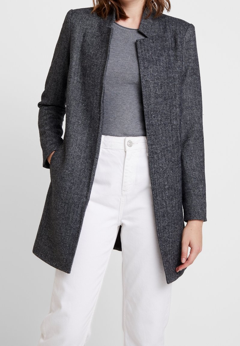 Femme portant un manteau en chevrons gris foncé sur un haut côtelé gris et un pantalon blanc taille haute, debout avec une main dans la poche.