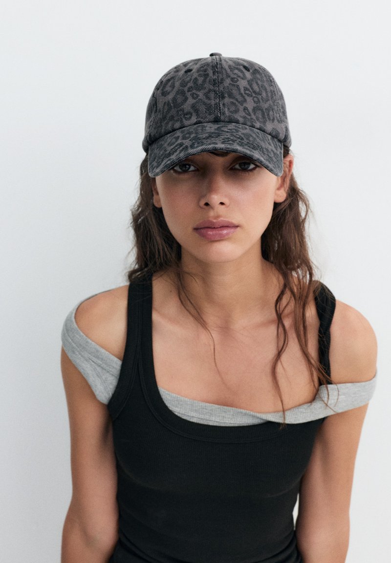 Jeune femme aux longs cheveux foncés portant une casquette grise à imprimé léopard et des hauts superposés noirs et gris, épaules dénudées, sur un fond uni.
