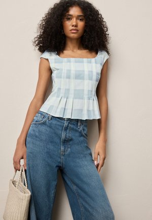 Vrouw met krullend haar die een mouwloos blauw-wit geruit topje en hooggetailleerde blauwe jeans draagt en een gevlochten beige handtas vasthoudt.