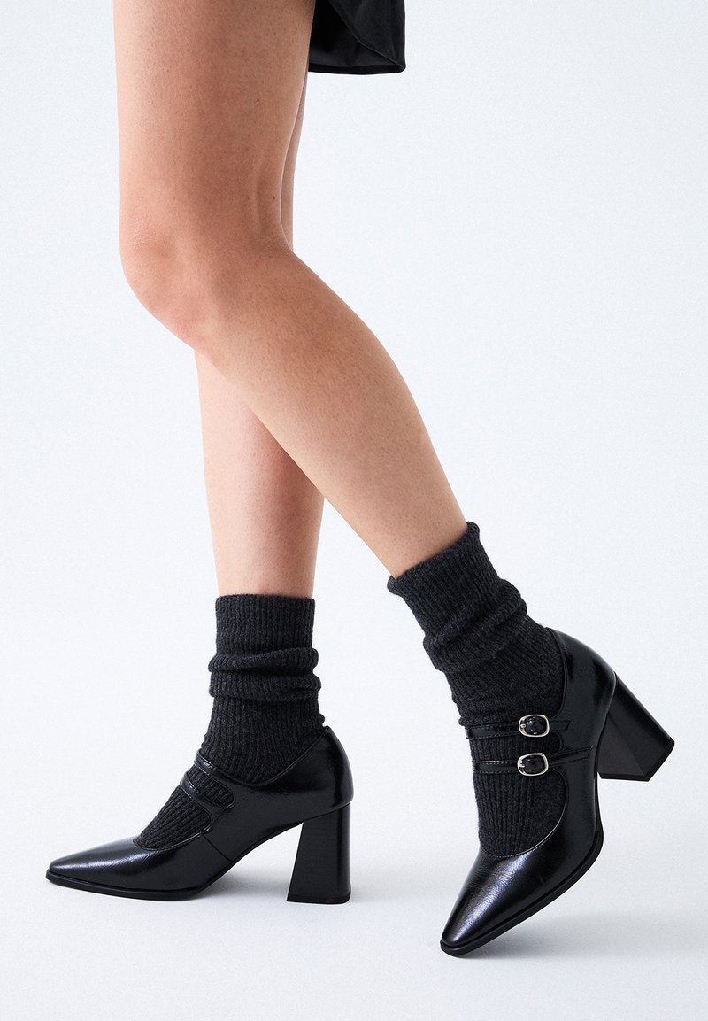 Zapatos de tacón de charol negro con punta afilada, que presentan dos detalles de hebilla y calcetines de carbón acanalados, creando una textura en capas.