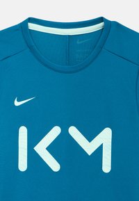 Camisola atlética azul Nike Dri-FIT com decote redondo, apresentando um grande logótipo branco "KM" com suaves detalhes às riscas. Textura suave.