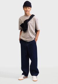 Light gray t-shirt with text, navy oversized jeans with yellow stitching, and white sneakers with black stripes; black beanie and sweatshirt draped.