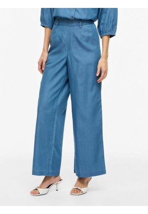 Femme portant un pantalon large bleu et un chemisier bleu assorti à des sandales blanches à talons ouvertes, debout devant un fond uni.