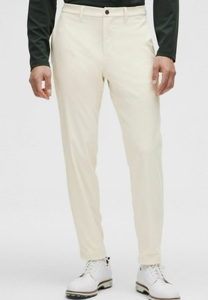 Pantaloni color crema a taglio slim con tasche frontali, chiusura con un solo bottone e una leggera lucentezza, abbinati a scarpe bianche.