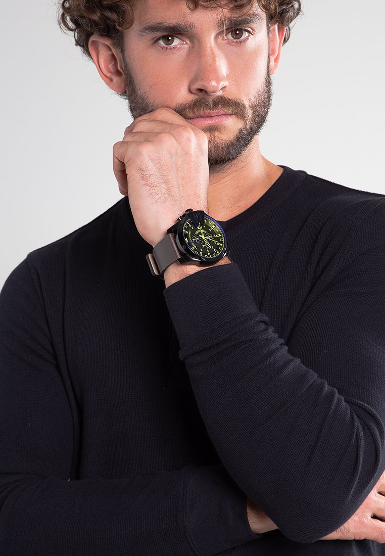 Reloj de pulsera negro con acentos amarillos, con una esfera redonda y una correa texturizada. Llevado en una muñeca con una camiseta negra de manga larga ajustada.