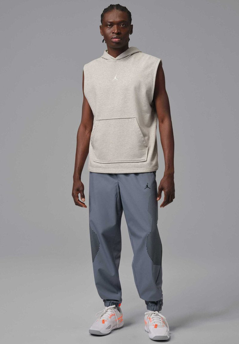 Jasnoszara bezrękawnikowa bluza z kieszenią z przodu; do niej ciemnoszare joggery z kropkowanymi akcentami i elastycznymi mankietami. Białe sneakersy.