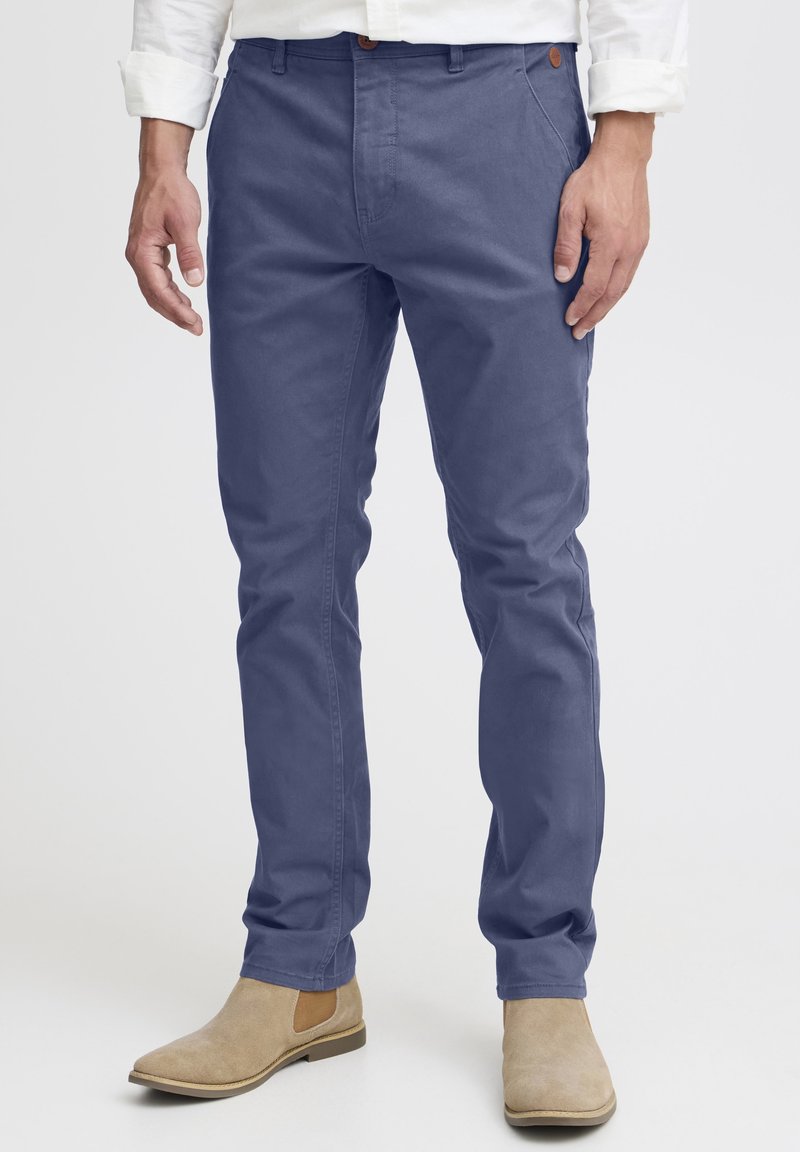 Homme portant un pantalon bleu marine slim, une chemise blanche et des bottines Chelsea en daim beige, debout devant un fond uni.
