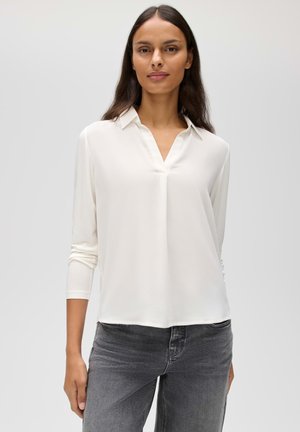 Frau mit langen dunklen Haaren, die eine weiße Bluse mit langen Ärmeln und graue Jeans trägt, steht vor einem schlichten hellen Hintergrund.