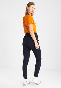 Oransje t-skjorte med trykk, kombinert med marineblå slim-fit bukser, med elastisk liv og lommer, brukt med hvite høye sneakers.