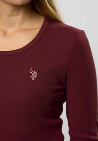 Tricou lung cu mânecă, de culoare burgundă, cu un decolteu rotund, având un mic logo brodat pe partea stângă. Material moale și elastic.