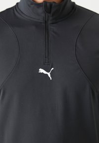 Juodas sportinis megztinis su aukštu apykakle ir ketvirčio užtrauktuku. Turintis glotnų audinį ir baltą Puma logotipą krūtinėje.