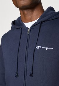Marineblaue Kapuzenjacke mit einem Frontreißverschluss, verstellbarem Kordelzug an der Kapuze und weißem "Champion"-Logo auf der Brust. Hergestellt aus weichem Stoff mit glatter Textur.