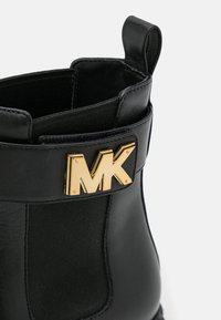 Bottine noire en cuir avec panneau latéral élastique et logo métallique doré MK sur la sangle près du bord supérieur.
