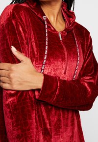 Hoodie en velours rouge avec fermeture éclair, motif crocodile texturé et cordons contrastés. Manches longues et coupe décontractée.