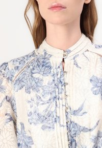 Bluză florală dintr-un material crem deschis, cu modele de flori albastre, guler înalt, detalii cu nasturi și pliseuri delicate pe partea din față.