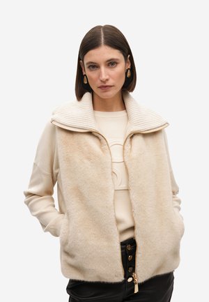 Chaleco de forro polar beige con cuello acanalado, cremallera dorada y bolsillos con cremallera. Lleva puesto un suéter color crema y pantalones negros. Textura suave.