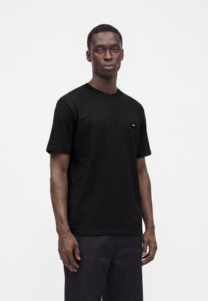 Homme à la peau foncée portant un t-shirt noir à manches courtes et un pantalon sombre, debout devant un fond clair uni avec une expression neutre.
