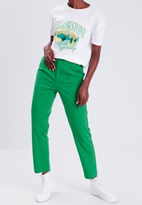 White t-shirt with a Yellowstone graphic, paired with green tailored pants and white high-top sneakers. Smooth textures and casual fit.
