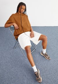 Hnědá oversize košile s krátkým rukávem, bílé šortky a hnědé sportovní boty s bílými ponožkami. Model sedí na kovové židli na modrém koberci.