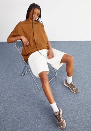 Brun oversized kortærmet skjorte, hvide shorts og brune atletiksko med hvide sokker. Model siddende på en metalstol mod et blåt tæppe.