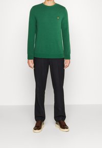 Zielony sweter na drutach z okrągłym dekoltem, ściągaczami na mankietach i u dołu. Dopasowane ciemne spodnie oraz brązowe trampki z kontrastową podeszwą.