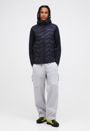 Persona in piedi con le mani nelle tasche, indossa una giacca trapuntata nera, pantaloni cargo grigio chiaro e scarpe giallo neon e nere.