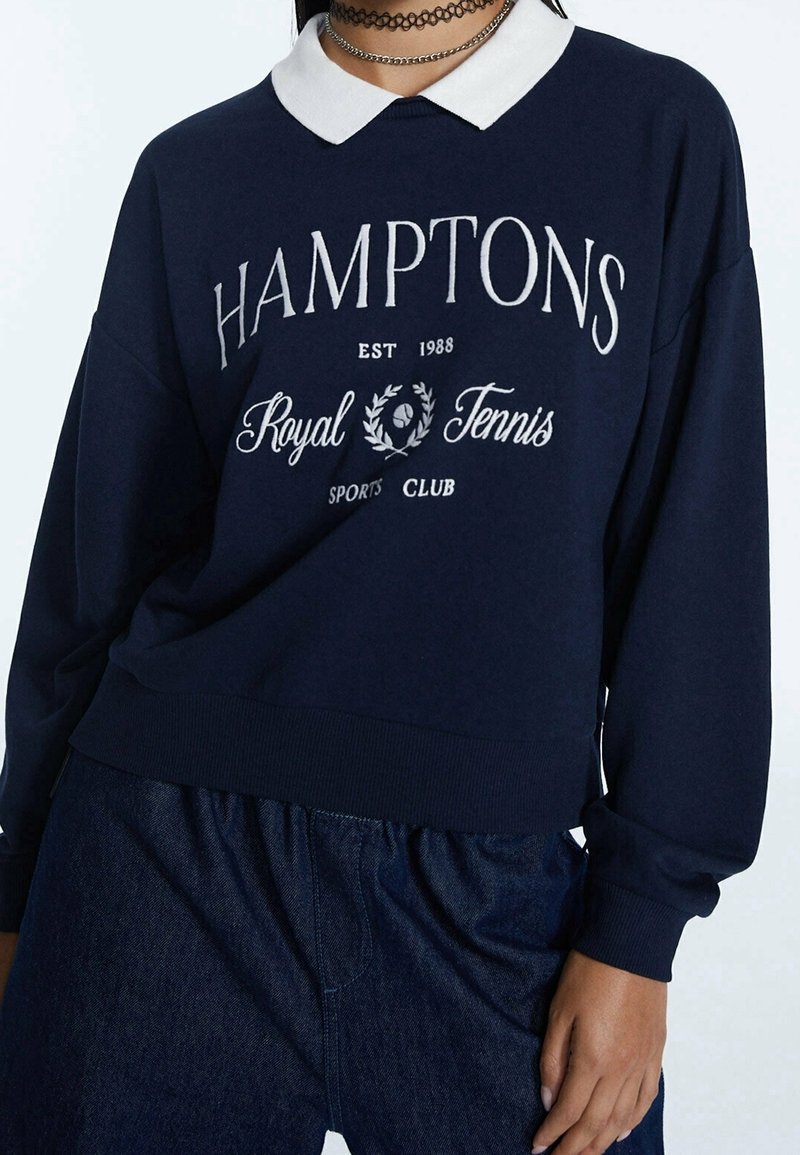 Sweat-shirt marine avec col blanc, présentant le texte "HAMPTONS Royal Tennis Sports Club" et des détails de design. Poignets et ourlet côtelés.