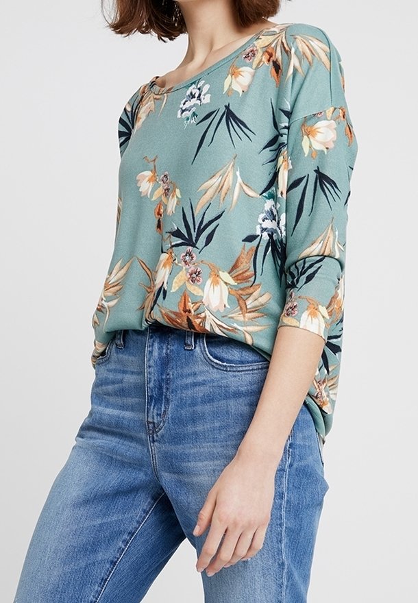 Langärmliges Blumenoberteil aus türkisgrünem Stoff, mit orangefarbenen, beigen und weißen Blumen, kombiniert mit hellblauen Jeans. Glatter und lockerer Schnitt.