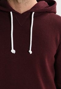 Bordeaux hoodie met een witte koord, met een V-vormige halslijn en textuurstof. Het ontwerp is eenvoudig en casual.
