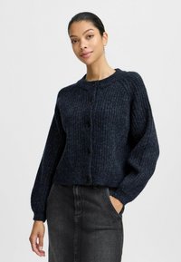 Tmavě modrý pletený cardigan s žebrovanou strukturou, kulatým výstřihem a předními knoflíky, v kombinaci s tmavými džíny sukně s bočním prošíváním.