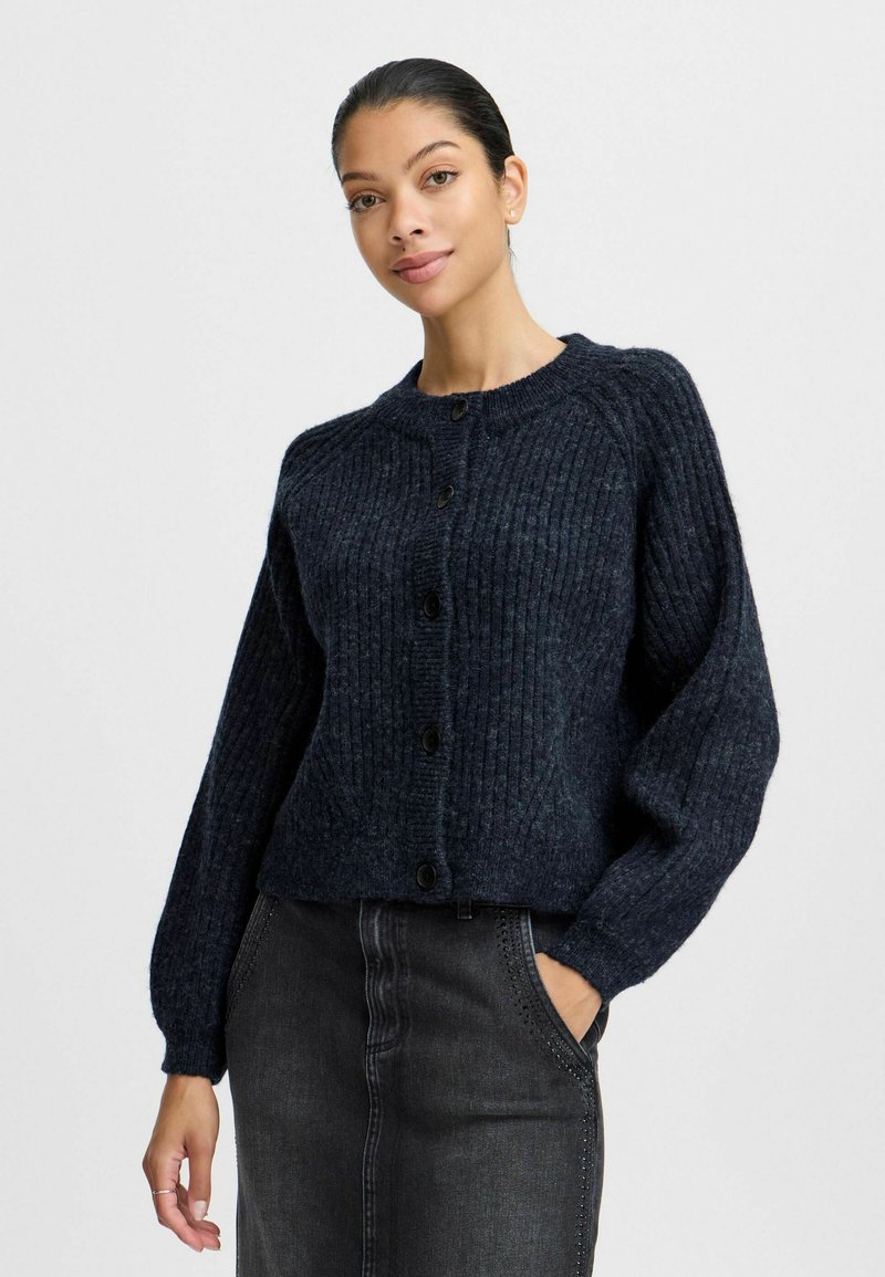 Tmavě modrý pletený cardigan s žebrovanou strukturou, kulatým výstřihem a předními knoflíky, v kombinaci s tmavými džíny sukně s bočním prošíváním.