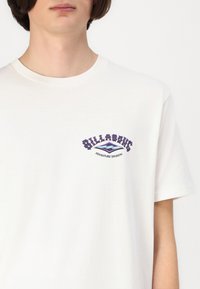 Hvit bomull T-skjorte med rund hals. Har grafisk tekst i lilla og blå farge "BILLABONG ADVENTURE DIVISION" på fronten.