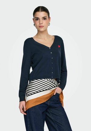 Ung kvinde med bagglattet hår iført navy cardigan med rød hjerteemblem, stribet tørklæde og mørke jeans, poserer med hånden i lommen.