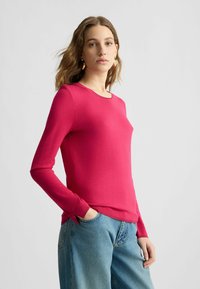 Fuchsia Langarmshirt aus einem weichen Strickstoff, mit rundem Halsausschnitt und gerippten Bündchen, kombiniert mit lockeren blauen Jeans.