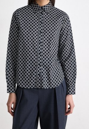 Kobieta ubrana w czarną koszulę zapinaną na guziki z białymi geometrycznymi krzyżykowymi wzorami, połączoną z wysokim stanem ciemnoszarymi plisowanymi spodniami.